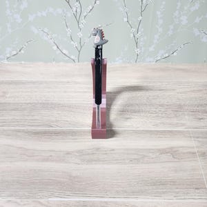 May include: A pen with a black ink cartridge and a decorative dinosaur topper. The pen is set in a pink pen holder. The background features a light green floral wallpaper.