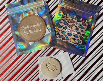 May include: Two holographic zip bags with dinosaur-themed designs and a round wooden coaster that says "Buttreraptor Butterbeer". A small, round, light-colored object with a dinosaur outline is also present.