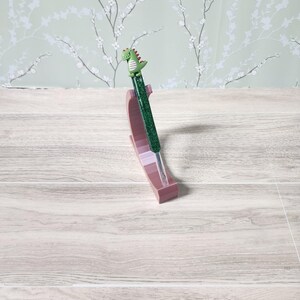 May include: A green glitter pen with a dinosaur figure on top is displayed in a pink pen holder. The pen has a green body and a small green dinosaur figure with red accents. The pen holder is a light pink colour.