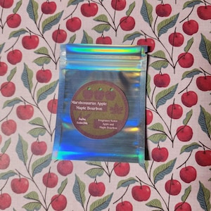 Könnte beinhalten: Ein holografischer, schillernder Beutel mit einem kreisförmigen Etikett mit der Aufschrift "Marshosaurus Apple Maple Bourbon". Das Etikett zeigt Apfelillustrationen und Duftnoten. Der Beutel befindet sich vor einem Hintergrund mit Kirschblütenmuster.