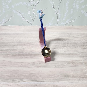 May include: A decorative spoon with a blue glitter handle and a small blue dinosaur figure on top. The spoon rests in a pink stand, with a gold-coloured spoon bowl.