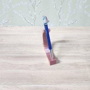 May include: A blue glitter pen, topped with a dinosaur figurine, displayed in a pink pen holder. The pen holder sits on a light wood-effect surface, with floral wallpaper in the background.