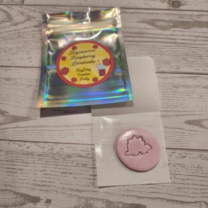 May include: A holographic pouch with a label reading "Rajasaurus Raspberry Lemonade" and a pink, circular creation with a dinosaur imprint. The items are on a wooden surface.