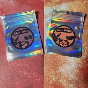 Może przedstawiać: Dwie holograficzne torebki, jedna z napisem "Dino Chills" z grafiką wiśni, a druga "Dino Drills" z grafiką dyni. Obie torebki zawierają "Tingling Creation Putty" i są zamknięte.