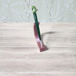 Może przedstawiać: Zielony długopis z brokatowym wykończeniem i małą zieloną figurką dinozaura na górze. Długopis jest prezentowany w różowym uchwycie na długopisy na jasnej powierzchni z fakturą drewna. Tło przedstawia kwiatową tapetę.