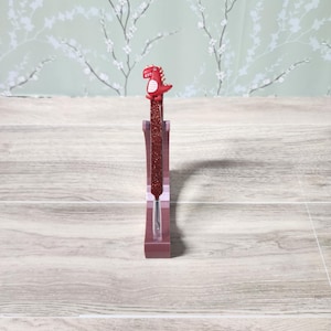 May include: A red glitter pen with a red dinosaur topper is displayed in a clear pen holder. The pen holder is a rectangular shape with a clear centre. The background is a light green floral pattern.