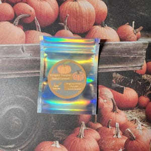 Puede incluir: Una bolsa holográfica con una etiqueta que dice "Eoraptor Pumpkin Salted Caramel" y "Notas de fragancia: Calabaza Caramelo Salado". La bolsa está colocada frente a un fondo de numerosas calabazas naranjas.