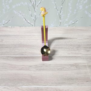 May include: A decorative spoon featuring a yellow dinosaur figure. The spoon has a gold-coloured bowl and a yellow handle. It rests in a purple holder. The background shows floral wallpaper.