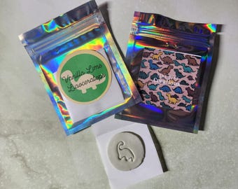 May include: Two small silver holographic bags with a white sticker on each. The sticker on the left bag says "Vanilla Lime Lioaceratops". The sticker on the right bag has a pink background with a pattern of colorful dinosaurs and the text "The Tiny Creations". A white paper with a gray dinosaur clay imprint is in the center of the image.