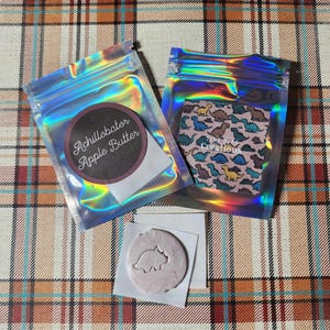 May include: Two iridescent zip-lock bags, one labelled "Achillobator Apple Butter" and the other with a dinosaur pattern. A small, pink, circular item with a dinosaur cutout rests on a white square.