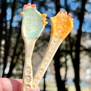May include: Two ceramic chicken-shaped spoons with floral designs. One spoon is light blue with white polka dots and the other is yellow with white polka dots.