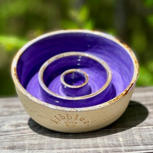 May include: Purple ceramic slow feeder bowl with a white rim and the word "Kibbles" inscribed on the bottom. The bowl has three concentric circles.