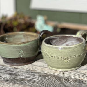 May include: Two handmade ceramic mugs. One is sage green with a brown base and a tree design. The other is light green with the words "merry yee merry" and snowflake designs. Both have dark brown handles.