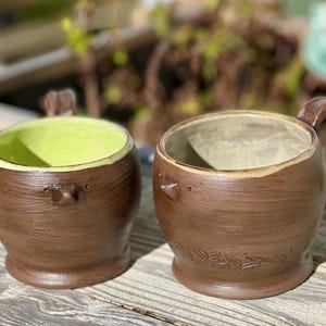 May include: Two handmade ceramic mugs. The mugs are brown with a textured finish. One mug has a bright green interior, while the other has a light beige interior. Both mugs have small, decorative protrusions and handles.