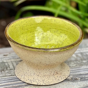 May include: A ceramic bowl with a pedestal base. The interior is a vibrant lime green, contrasting with the speckled beige exterior. The rim has a dark brown accent. The bowl is likely used for serving or display.