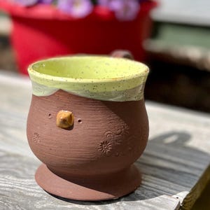 May include: A handmade ceramic mug with a brown exterior and a light green interior. The mug features a small, square, tan-colored embellishment and embossed floral designs. The rim is painted with a yellow band. The mug sits on a wooden surface.