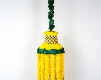 May include: A decorative hanging ornament featuring a green stem with spherical accents, a gold-colored bell-shaped top, and cascading yellow floral strands. The ornament has a green and yellow floral band and gold-colored bells at the bottom.