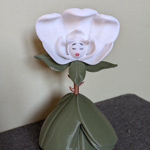 Peut inclure: Une fleur blanche avec un visage sculpté au centre. La fleur est sur une tige verte avec des feuilles vertes.