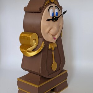 Pu&ograve; includere: Un orologio stampato in 3D di colore marrone e oro con un viso che ricorda un personaggio dei cartoni animati. L'orologio ha un pendolo e una lancetta nera che punta al numero 12.