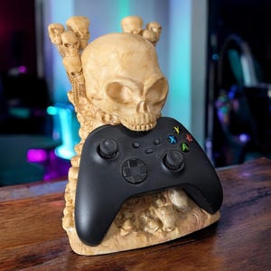 Puede incluir: Un mando Xbox negro se encuentra en un soporte con forma de cráneo hecho de resina beige. El soporte presenta múltiples cráneos más pequeños y detalles óseos.