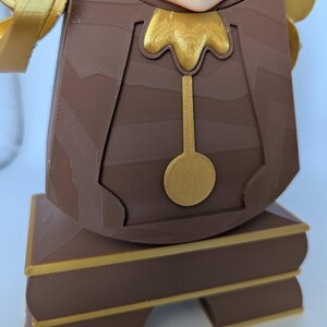 Pu&ograve; includere: Una statuetta marrone e dorata di un orologio con un viso umano. L'orologio ha un pendolo dorato e una base dorata.
