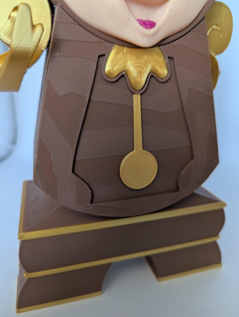 Pu&ograve; includere: Una statuetta marrone e dorata di un orologio con un viso umano. L'orologio ha un pendolo dorato e una base dorata.