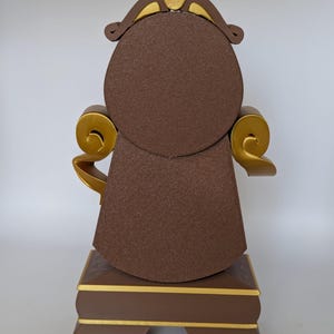 Pu&ograve; includere: Un orologio marrone e dorato a forma di sedia con un quadrante rotondo. L'orologio ha una corona dorata in cima e dettagli dorati sui lati. L'orologio &egrave; su una superficie bianca.