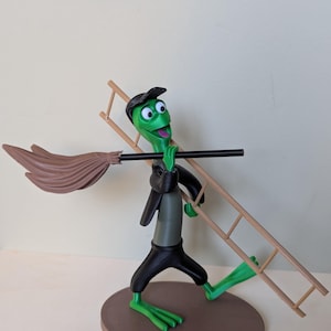 Peut inclure: Figurine de grenouille verte de dessin animé portant une veste, un pantalon et une casquette noirs, portant une échelle et un balai. La grenouille a un large sourire et se tient sur une base circulaire marron. Le balai est marron et l'échelle est beige.