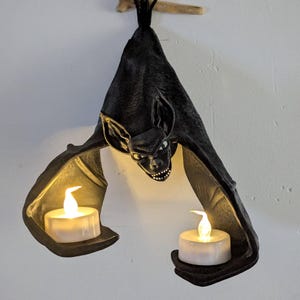 May include: A black bat-shaped wall sconce with a menacing face and outstretched wings. The bat holds two battery-operated candles in its wings. The sconce is mounted on a wooden branch, creating a spooky Halloween decoration.