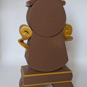 Pu&ograve; includere: Un orologio marrone e dorato con un quadrante rotondo e dettagli decorativi. L'orologio &egrave; in piedi su una base con tre gambe.