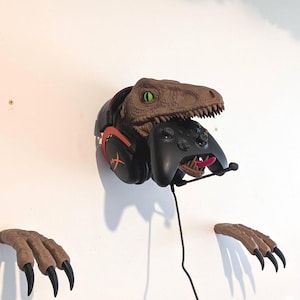 Könnte beinhalten: Eine braune Dinosaurierkopf-Wandhalterung, die einen schwarzen Gaming-Controller und ein Headset mit orangefarbenen Akzenten hält. Das Maul des Dinosauriers hält den Controller und seine Zunge ist rosa. Die Wandhalterung enthält auch zwei Dinosaurierkrallen.