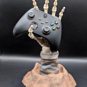 May include: A black Xbox controller is held by a skeletal hand, mounted on a brown, textured base. The controller features coloured buttons and a central directional pad. The base has a layered, earthy appearance.