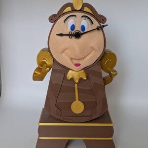 Pu&ograve; includere: Una statuetta di orologio in legno marrone e dorato di Cogsworth, dal film Disney La Bella e la Bestia. L'orologio ha un viso sorridente con occhi blu e una lancetta nera. L'orologio &egrave; in piedi su una base marrone con dettagli dorati.