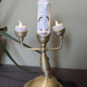 Op de afbeelding: Een gouden kandelaar met een witte kaars die een gezicht heeft. De kaars wordt verlicht met een klein lampje. De kandelaar heeft twee armen, elk met een klein lampje aan het uiteinde.