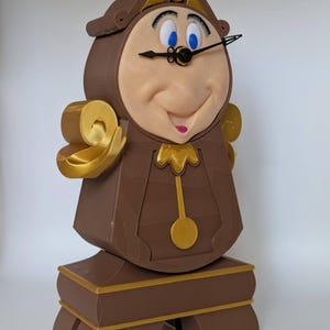 Pu&ograve; includere: Una statuina di Cogsworth, l'orologio di La Bella e la Bestia della Disney, stampata in 3D in marrone e oro. La statuina ha un viso dettagliato con occhi azzurri e una bocca sorridente. Il quadrante dell'orologio ha lancette nere e un pendolo dorato.