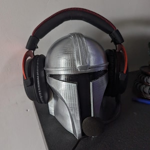 Puede incluir: Un casco mandaloriano de impresión 3D plateado con una visera negra, utilizado como soporte para auriculares. Un auricular negro con detalles rojos descansa sobre el casco.