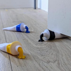 Puede incluir: Tres originales topes para puertas con forma de tubos de pintura, cada uno con un color diferente saliendo. Uno tiene pintura azul, otro amarilla y el tercero negra. Los tubos son blancos con tapas de colores, sobre un suelo de madera.