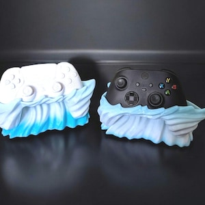 Peut inclure: Deux manettes de jeu vidéo, l'une blanche et l'autre noire, sont présentées dans des supports en forme de vague bleu et blanc.
