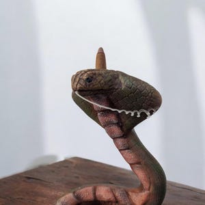 Op de afbeelding: Een keramiek cobra-beeldje met een groen en bruin gepatroneerd lichaam. De cobra staat in een verhoogde positie met zijn kap uitgespreid. Het beeldje staat op een houten oppervlak.
