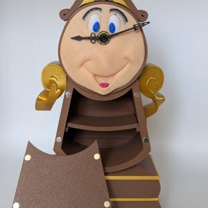 Pu&ograve; includere: Un orologio marrone e dorato con un viso umano, occhi blu e una bocca rossa. L'orologio &egrave; aperto e ha degli scaffali all'interno. L'orologio &egrave; un personaggio del film La Bella e la Bestia.
