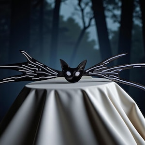 Puede incluir: Una diadema de Halloween en blanco y negro con un diseño de murciélago. La diadema tiene una cara de gato con ojos blancos y largas extensiones en forma de ala con rayas blancas. Se muestra sobre una superficie blanca contra un fondo oscuro y borroso de bosque.