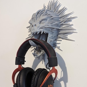 May include: A 3D printed grey monster head with spiky hair and an open mouth, used as a headphone holder. A pair of black headphones with orange accents are hanging from the monster's mouth.