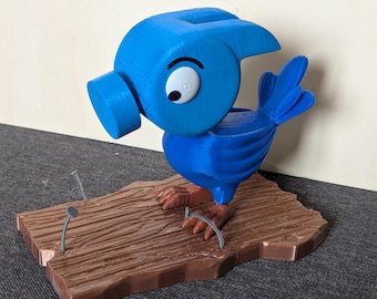 May include: A blue 3D printed bird figurine holding a hammer, standing on a brown wooden base with two nails. The bird has a large, white eye and a whimsical expression.