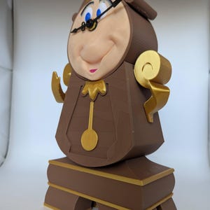 Pu&ograve; includere: Una statuetta in 3D stampata in marrone e oro di Cogsworth, il personaggio dell'orologio di La Bella e la Bestia della Disney. Ha un viso grande e rotondo con un quadrante dell'orologio e lancette, e un ampio sorriso con i denti. &Egrave; in piedi su una base con finiture dorate.