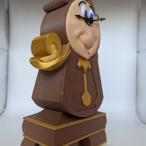 Pu&ograve; includere: Una statuetta di Cogsworth, il personaggio dell'orologio di La Bella e la Bestia della Disney, stampata in 3D in marrone e oro. La statuetta ha un viso dettagliato con occhi blu e una corona dorata. Il quadrante dell'orologio &egrave; visibile con lancette dorate.