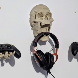 Puede incluir: Una configuración de juego montada en la pared con una calavera y dos manos esqueléticas que sostienen mandos de juego negros. Un par de auriculares negros y rojos cuelgan debajo de la calavera. La calavera y las manos son blanquecinas.