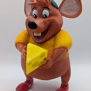 Puede incluir: Un ratón de dibujos animados marrón y naranja con un sombrero verde y una camisa amarilla sostiene un trozo de queso amarillo en sus manos.