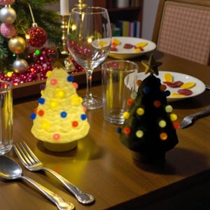 Peut inclure: Une table de Noël décorée avec un sapin et des ornements. Deux petits bougeoirs en forme d'arbre sont ornés de pompons colorés. Couverts, verres et assiettes avec des bonbons complètent la scène festive.