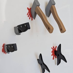 May include: A set of novelty wall hooks shaped like axes, knives, and throwing stars. The axes have grey blades and brown handles, the knives are black and grey, and the throwing stars are black. Red blood-like splatters are included.