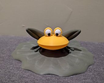 May include: A yellow frog figurine with large white eyes and a green lily pad base. The frog is sitting on the lily pad with its mouth open.